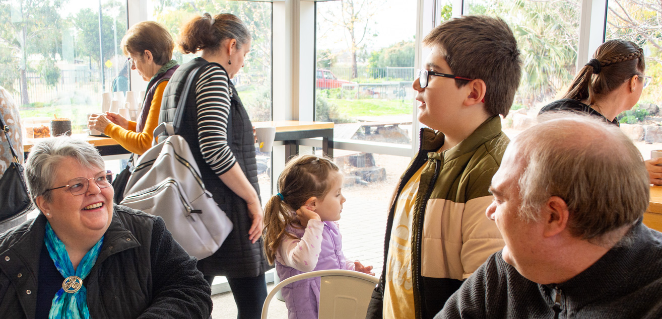 A group of people, both young and old gather together in the foyer. Some engaged in conversation, smiling and laughing together, as coffee is being poured in the background.