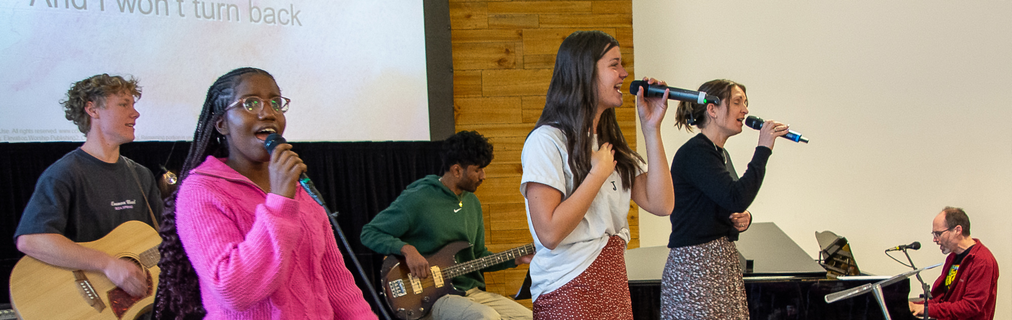 The worship team stands on the stage, microphones and instruments in hand as the sing together praising God.