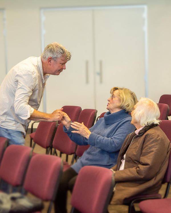 Pastor Peter Riggs reaches out with love and care and holds hands with one of two elderly ladies seated in the church.