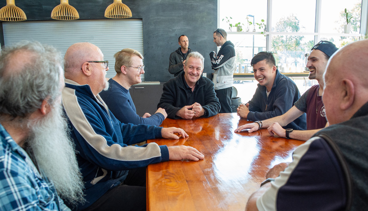 A gathering of men young and old, centered around a table, chatting, laughing and smiling together.