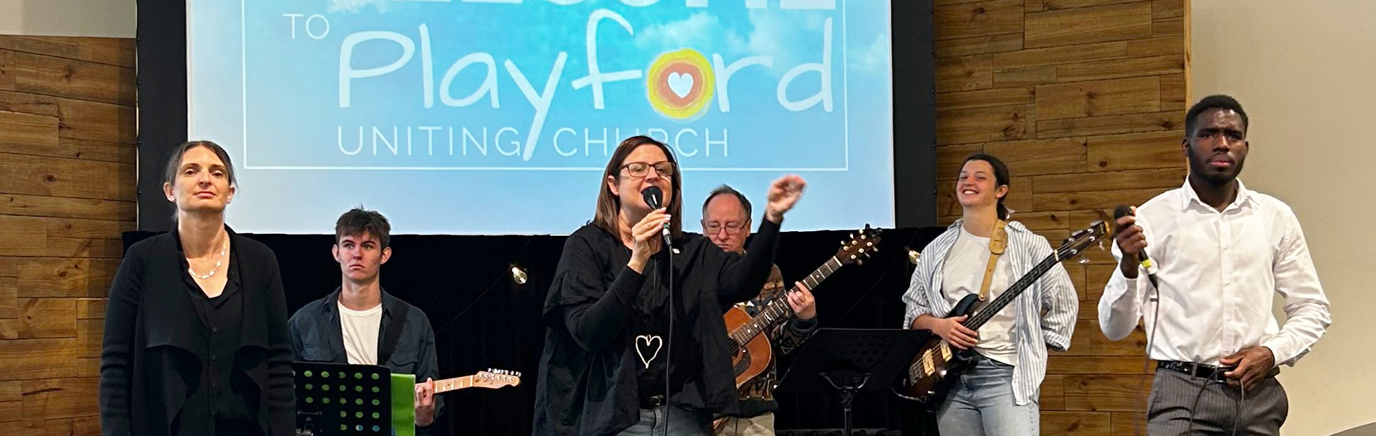 The church worship team, on the stage at the front of the building. Three people are signing and raising their hands worshiping God. Three others have guitars and are providing accompanying music.