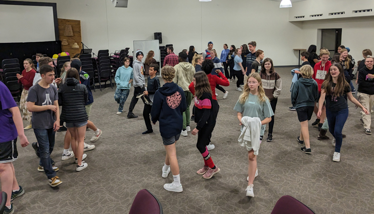 A large group of young people gathered together in the church building. They are all walking around circling each other, engaged in a game.